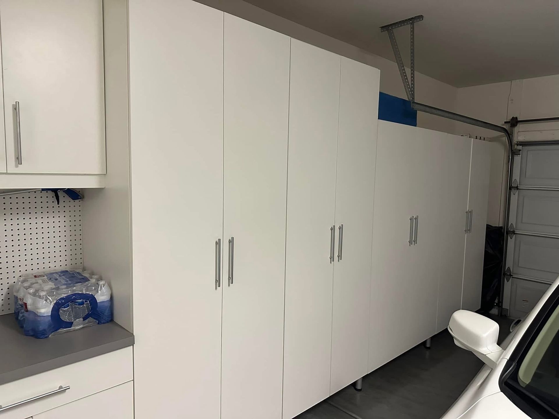 White storage cabinets and a counter in a garage, with a white chair and garage door track on the right.