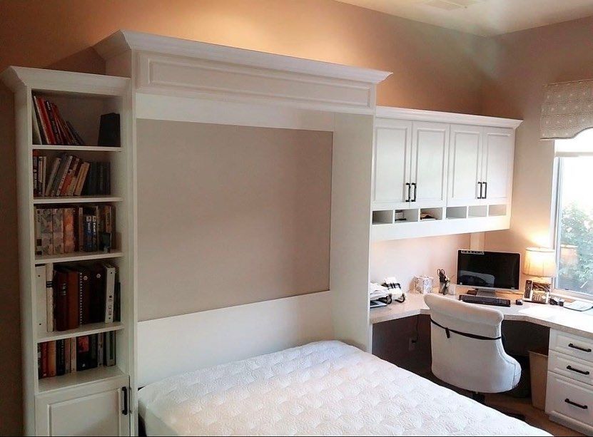 Bedroom office nook with built-in desk, white cabinets, bookshelves, and a Murphy bed under warm lighting