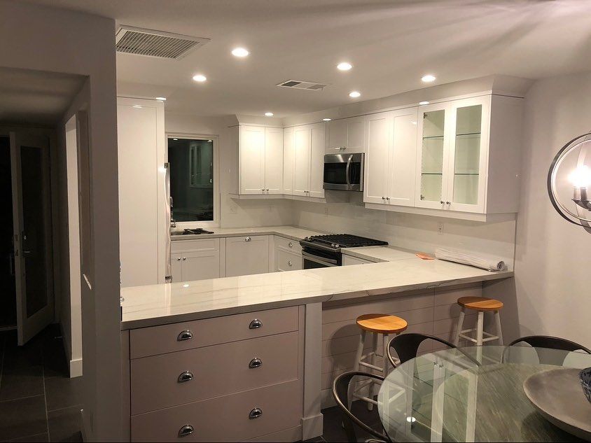 Modern white kitchen with island, glass dining table, and two wooden stools under recessed lights