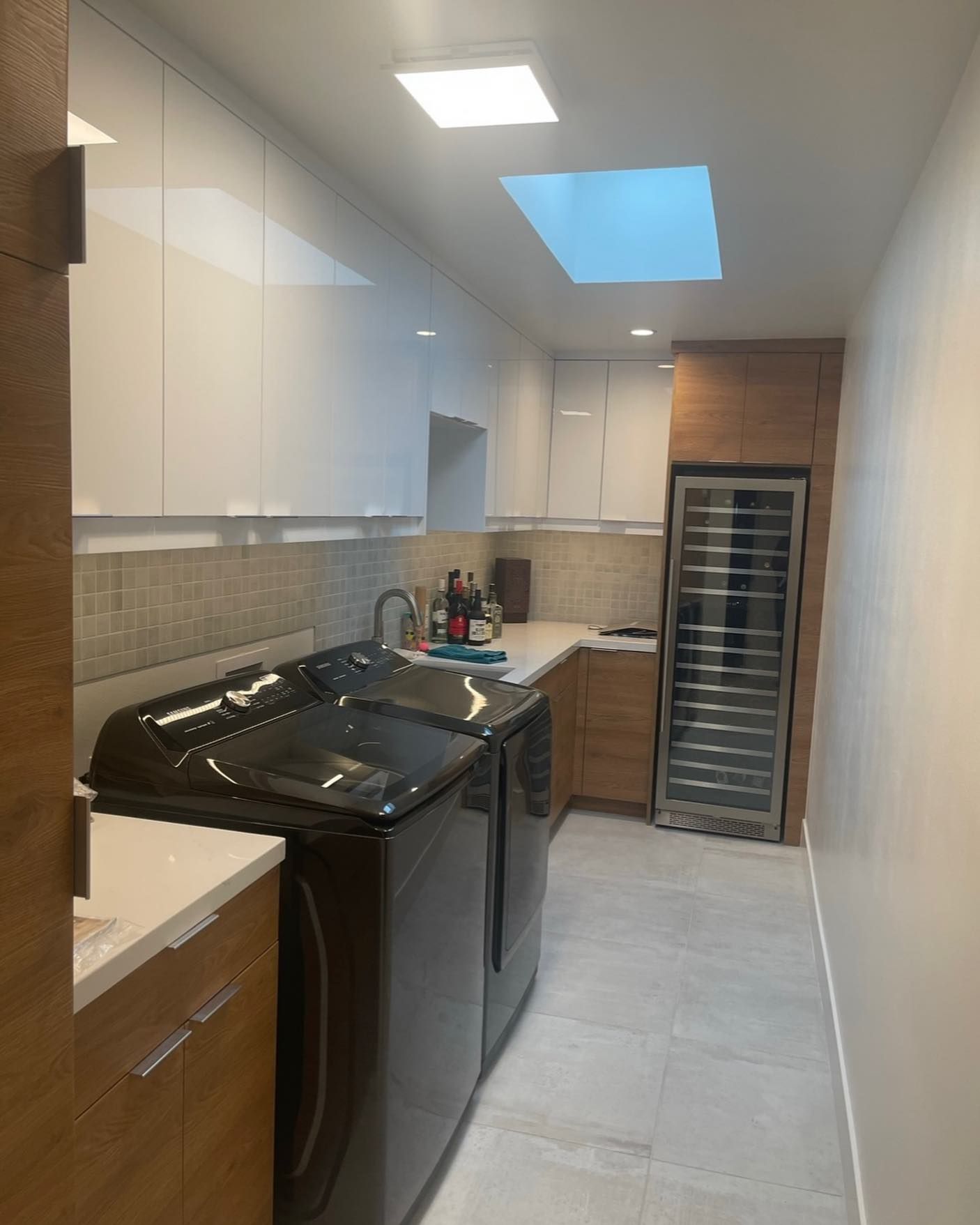 Narrow modern laundry room with washer, dryer, cabinets, and tiled floor under a skylight.