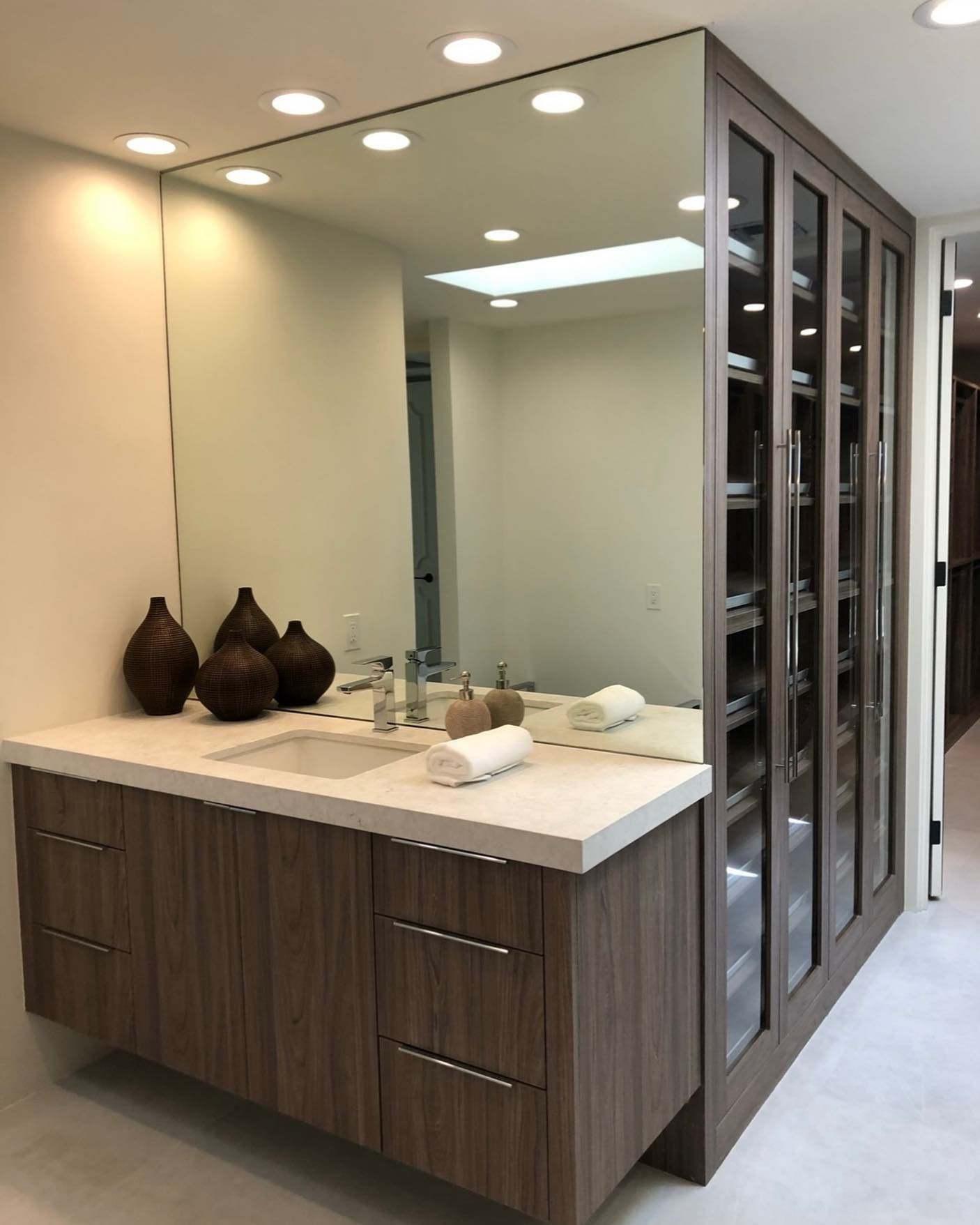 Modern bathroom vanity with large mirror, double sinks, wood cabinetry, and glass-front storage cabinets.