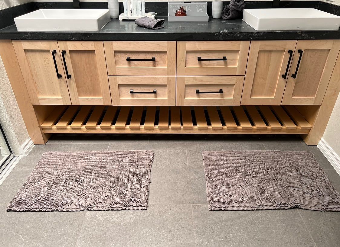 Modern wooden bathroom vanity with double sinks, drawers, and two gray bath mats on a tiled floor