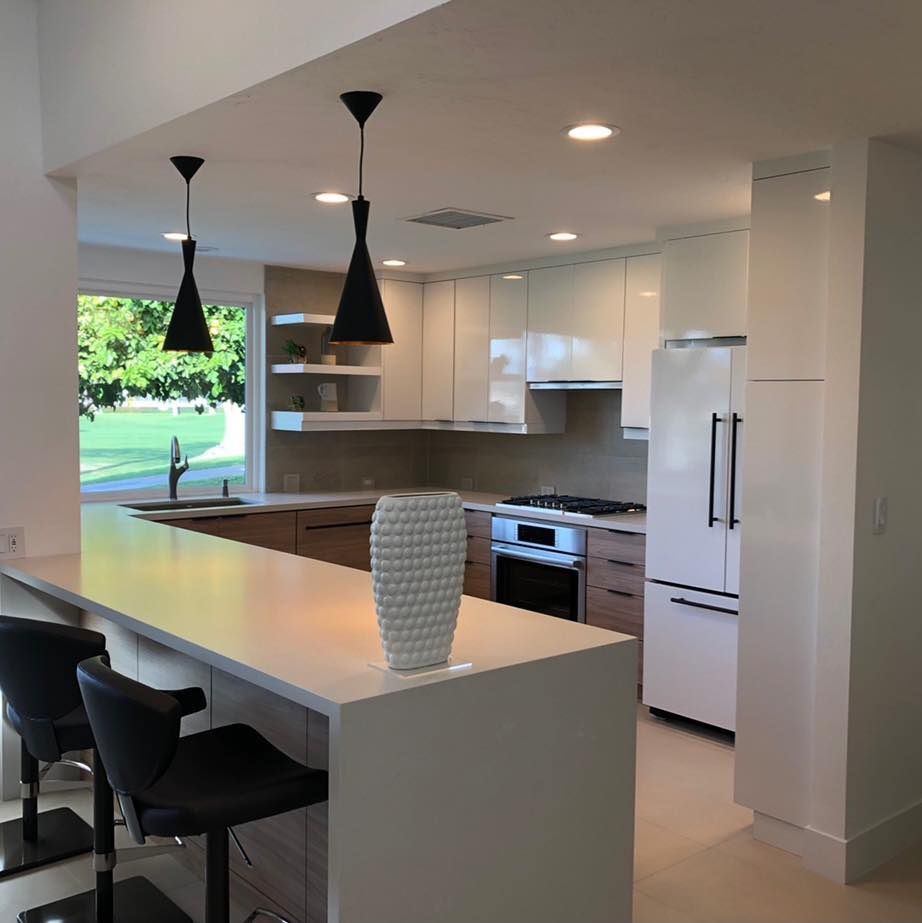 Modern white kitchen with island, black pendant lights, bar stools, and a window overlooking greenery