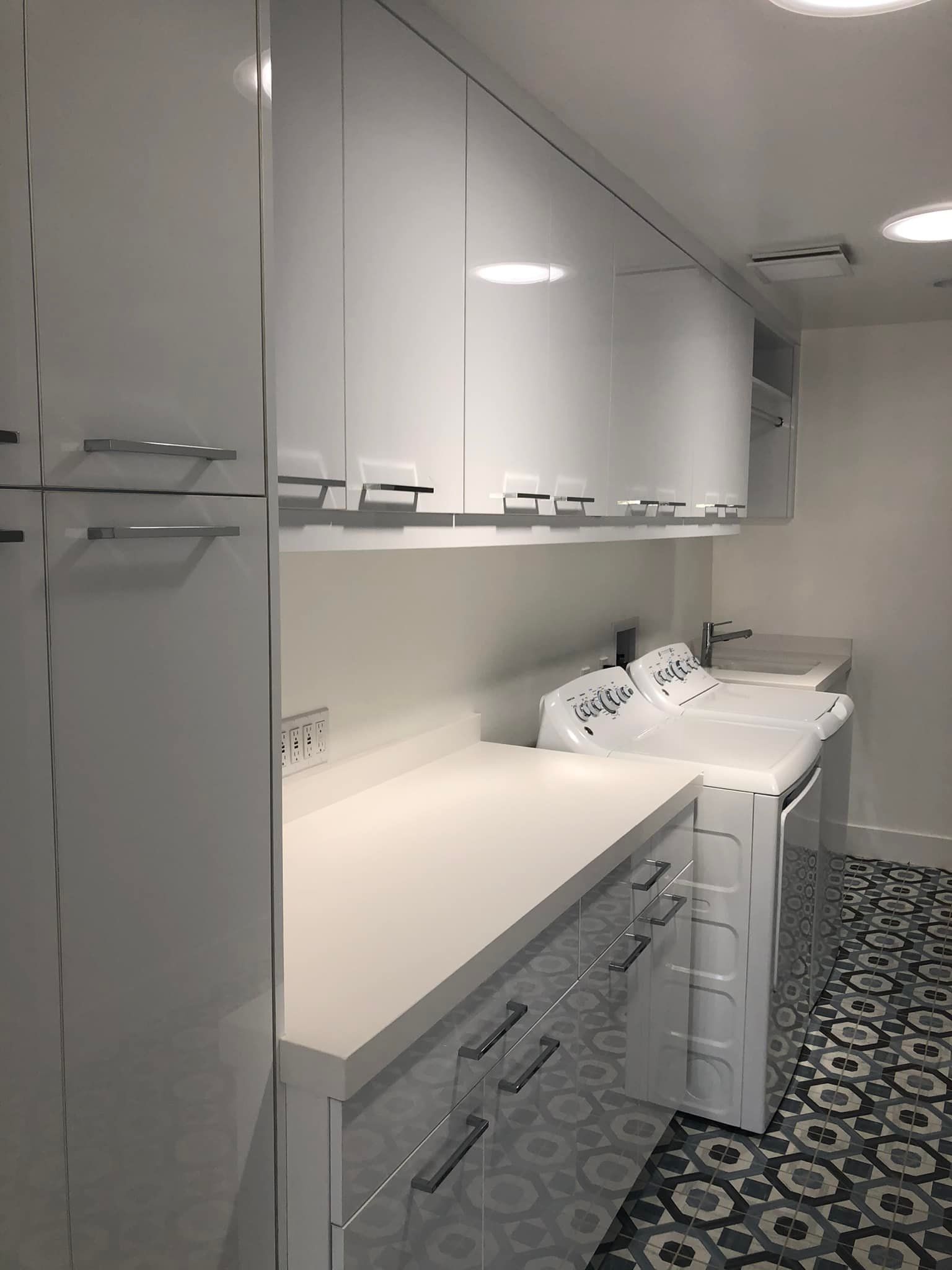 Modern white kitchen with glossy cabinets, long countertop, built-in sink, and patterned black-and-white floor.