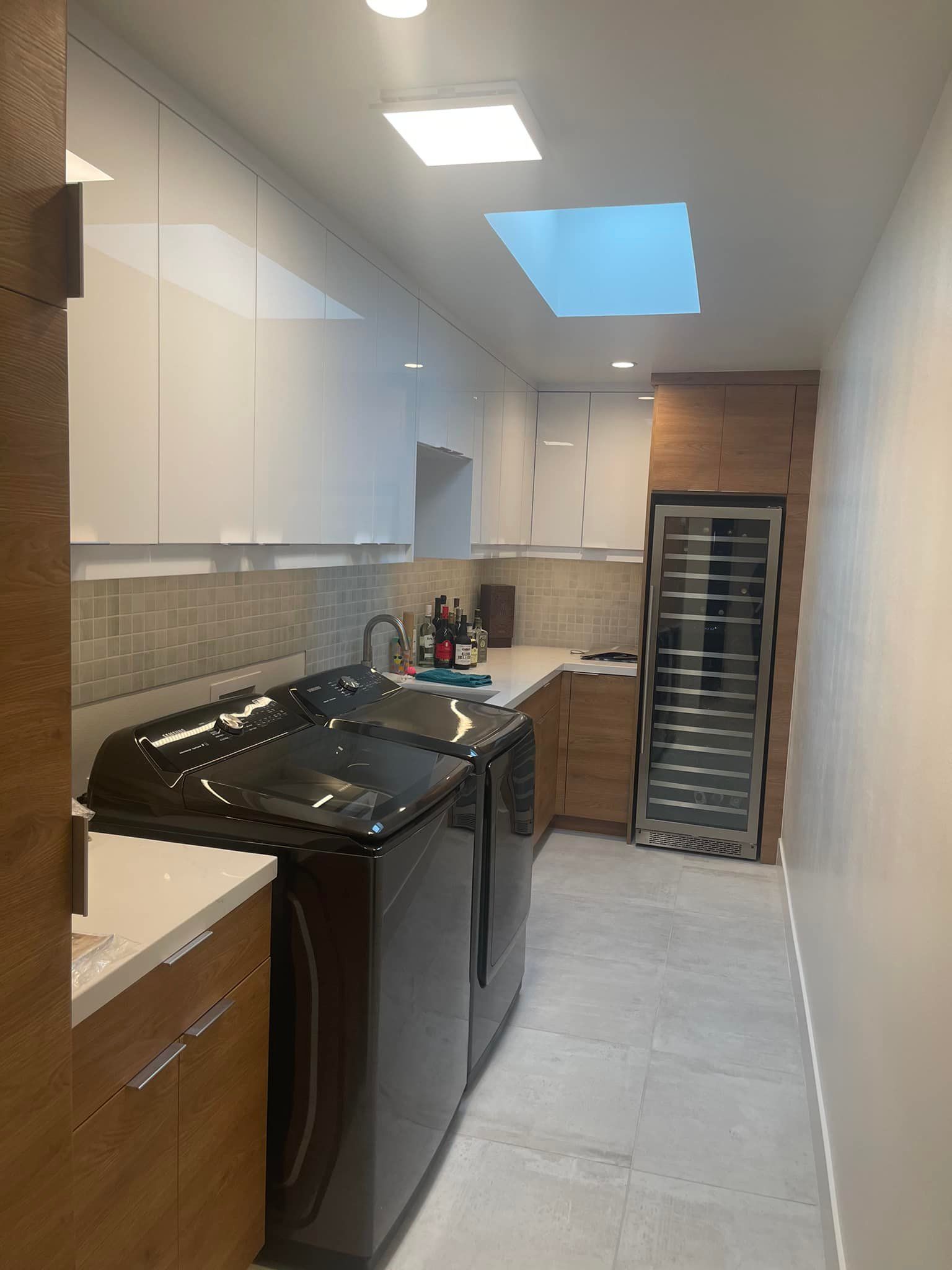 Narrow laundry room with black washer and dryer, white cabinets, tiled floor, and skylight.