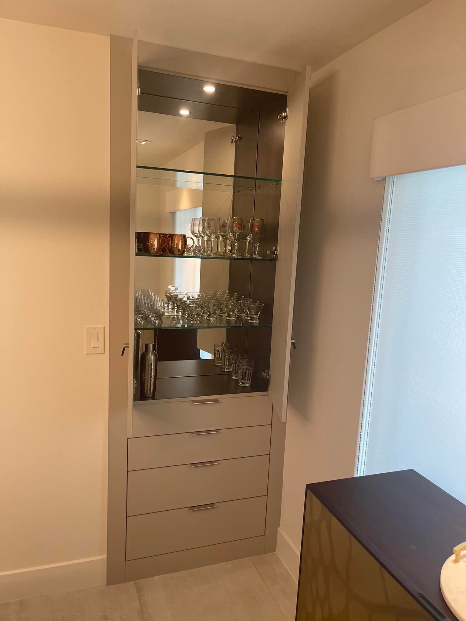 Built-in mirrored cabinet with glass shelves in a beige room beside a window and black-topped dresser