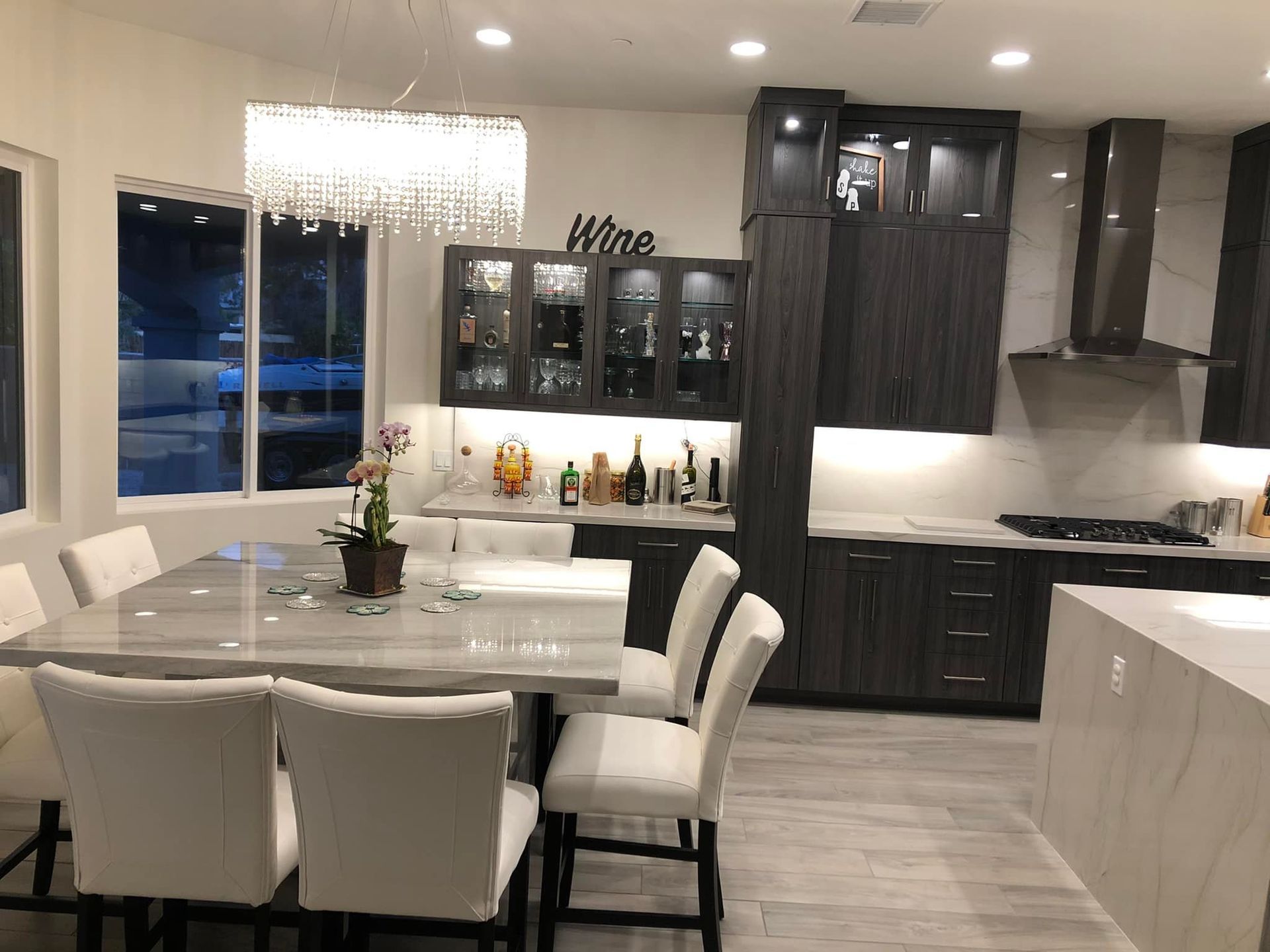 Modern kitchen and dining area with white chairs, dark cabinets, and a chandelier at night