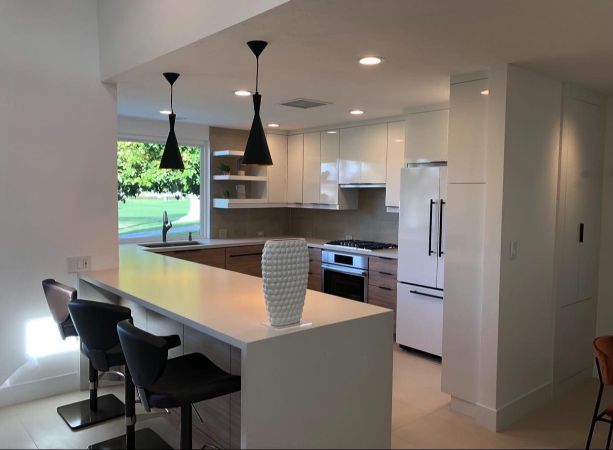 Modern white kitchen with island, black pendant lights, and stools, opening to a bright windowed dining area