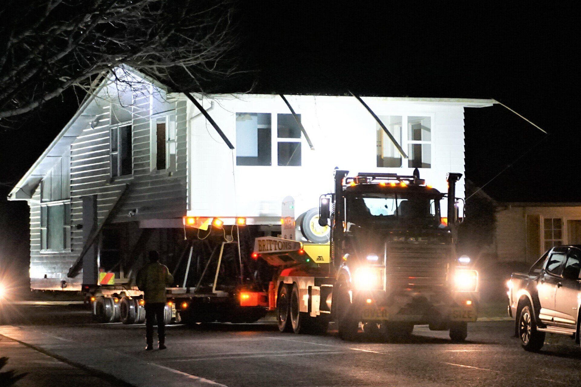 House being hauled by trucks at night on a dark road, headlights shining.