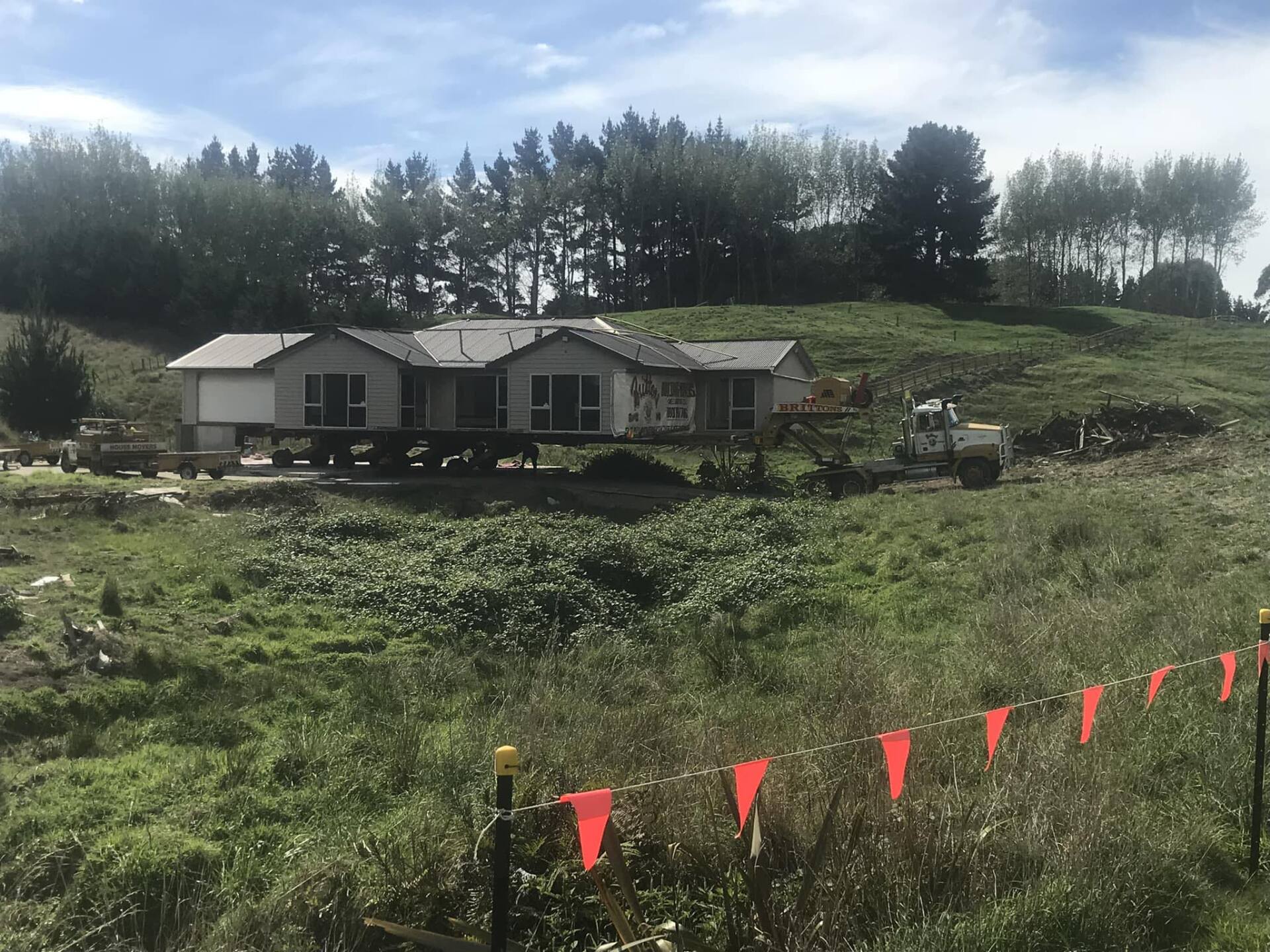 Single-story house in a grassy field under cloudy sky, with red flags in the foreground and trees behind.