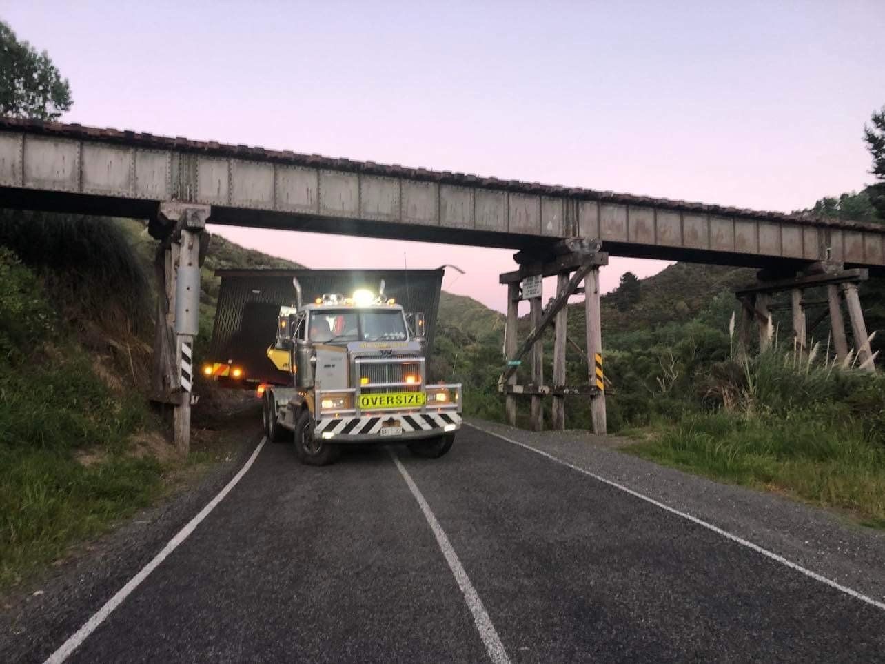 Truck driving under a low bridge on a rural road, with another vehicle behind it.