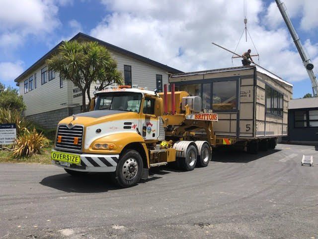 Yellow truck hauling a large glass-sided building module along a paved lot