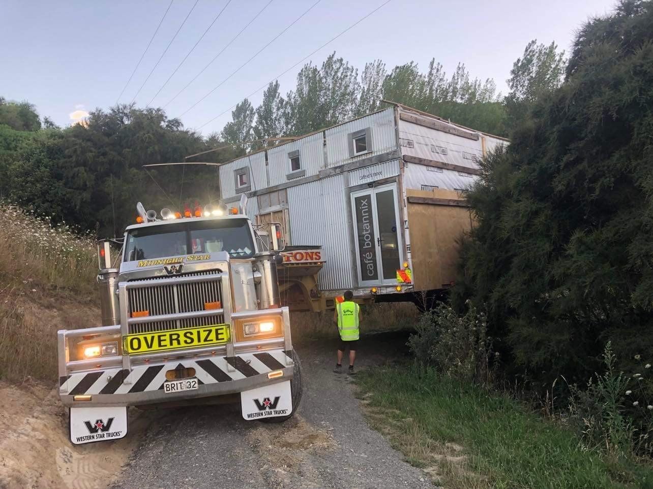 Tow truck hauling oversized white trailer through a narrow wooded dirt road