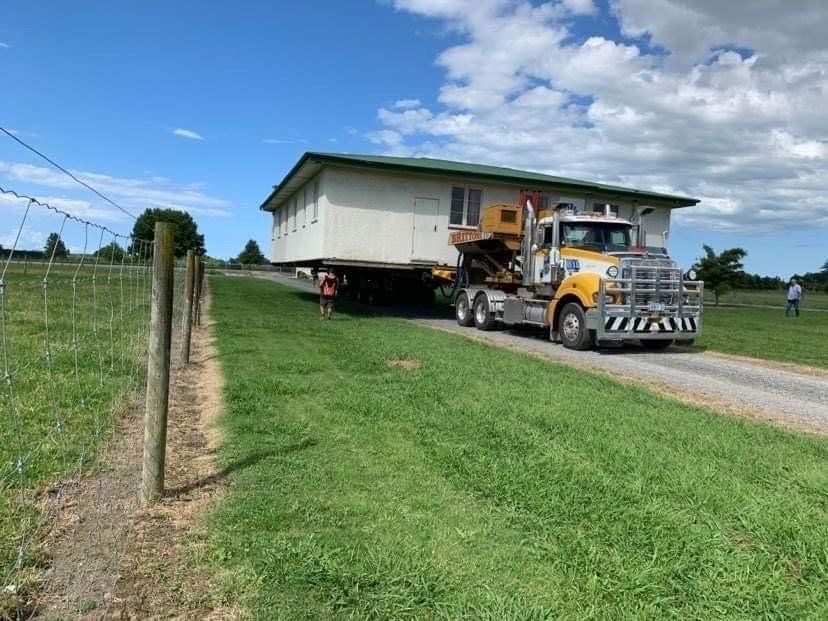Mobile home being hauled by a yellow truck on a rural road beside a grassy field under a blue sky