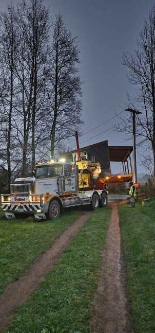 Fire truck parked on a grassy track at dusk, with emergency lights on near a small building and bare trees.