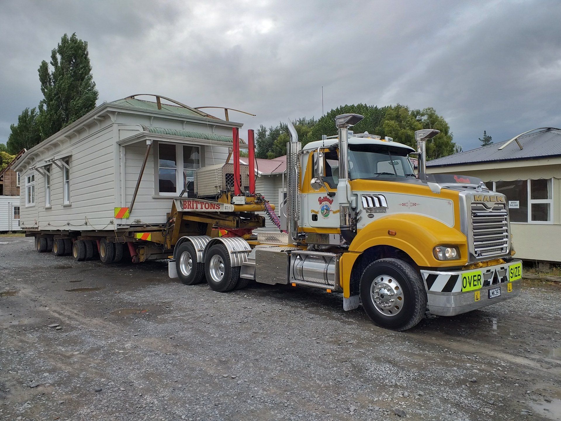 Yellow tow truck hauling a white house on a flatbed trailer in a gravel lot