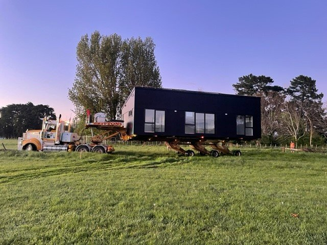 Black modular house being transported across a grassy field by a truck and trailer under a clear sky