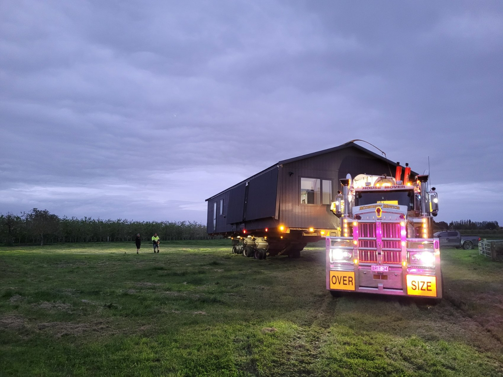 Colorful semi truck parked beside a building in a grassy field at dusk under a cloudy sky