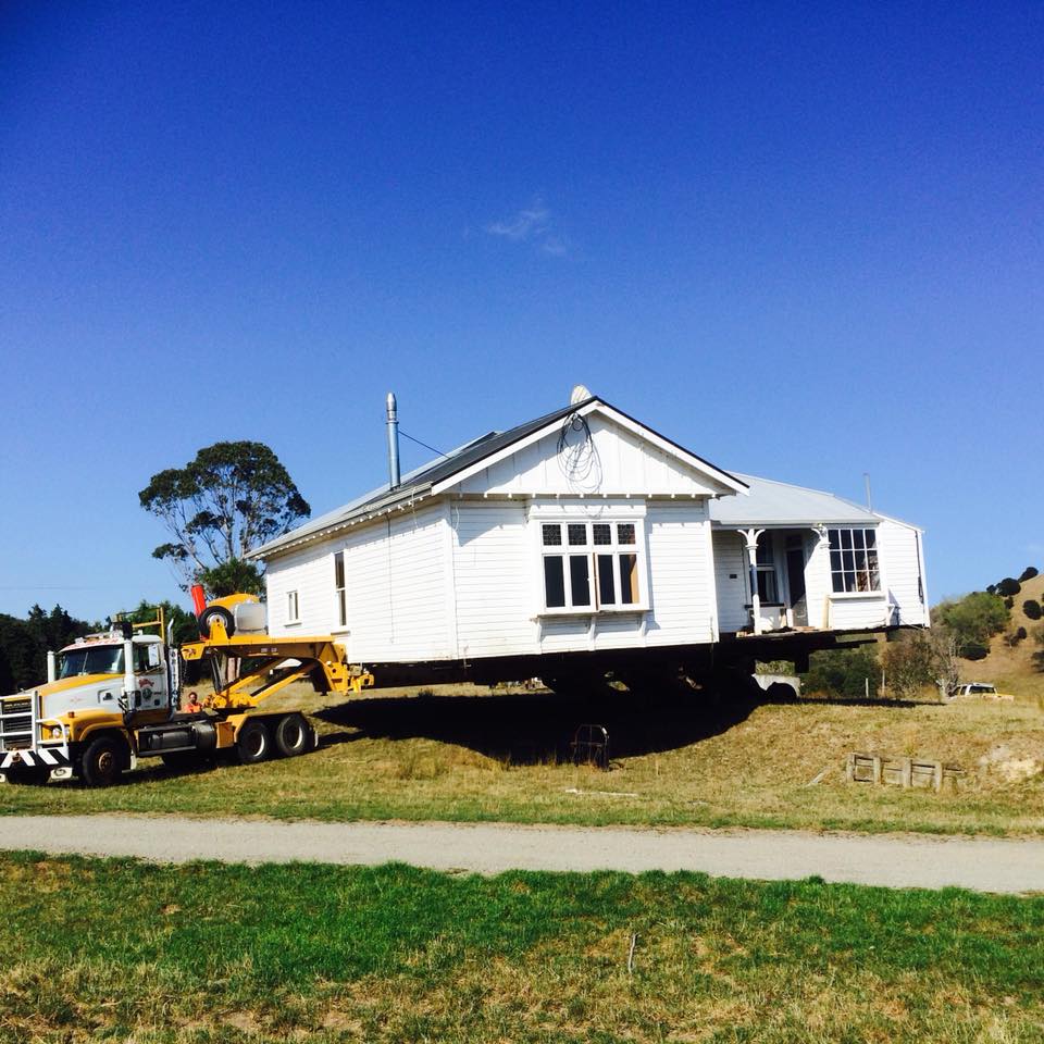 House being lifted by a yellow truck on a grassy lot under a clear blue sky