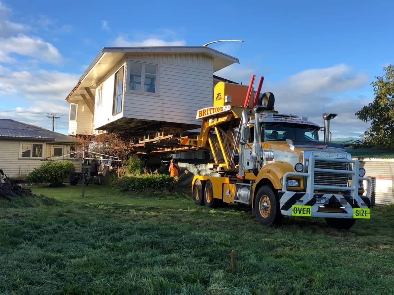 House being lifted by a crane onto a truck in a grassy yard under a blue sky