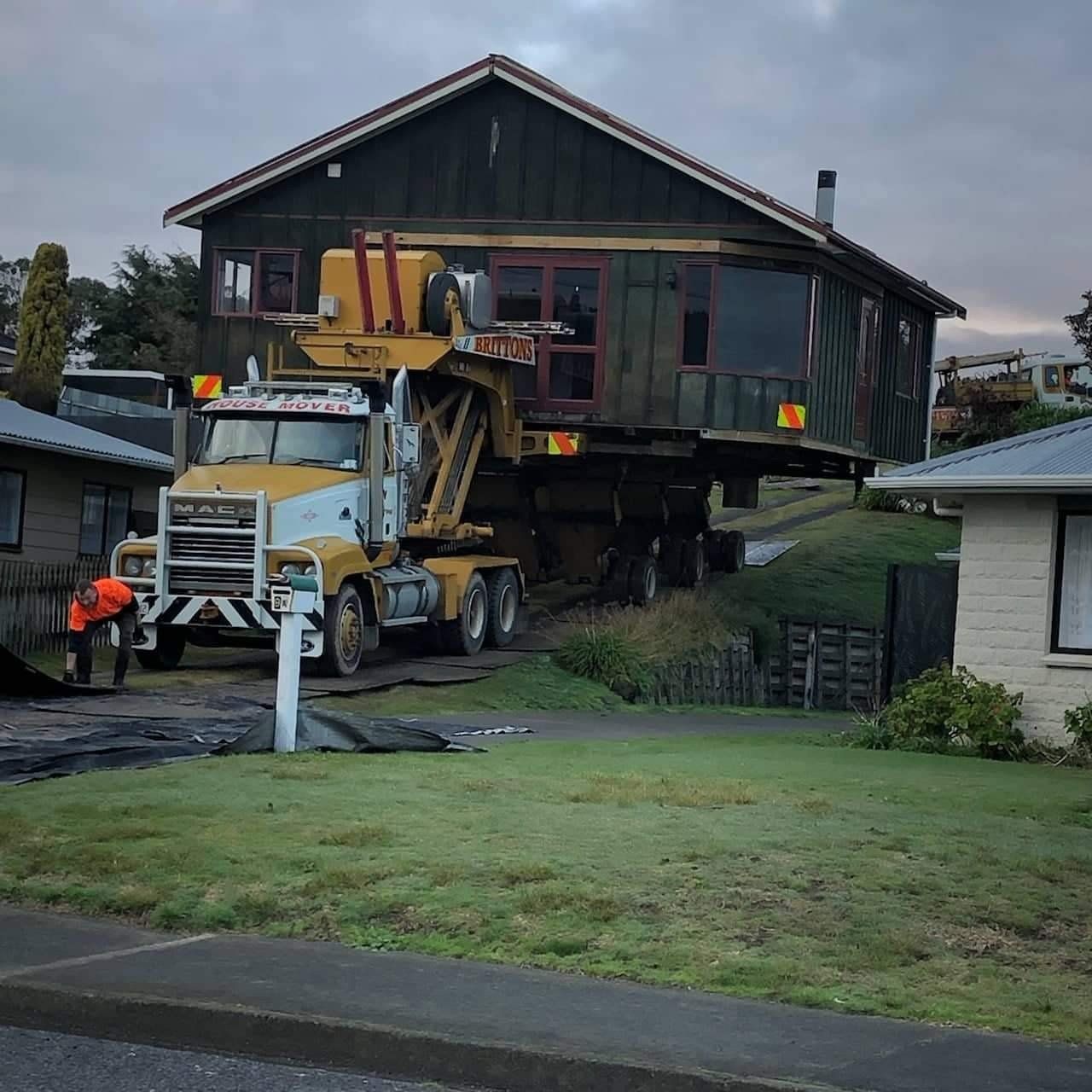 Yellow crane truck lifting a house on a residential street, with the house elevated above the trailer.