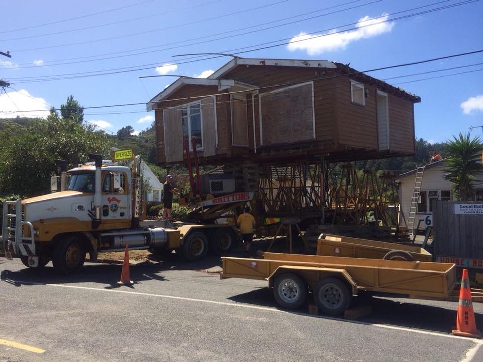 House being lifted by a crane onto a truck in a residential street, with traffic cones nearby