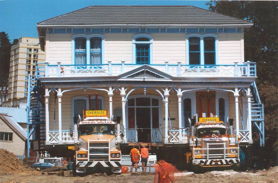 Two construction trucks parked in front of a white Victorian-style house under renovation