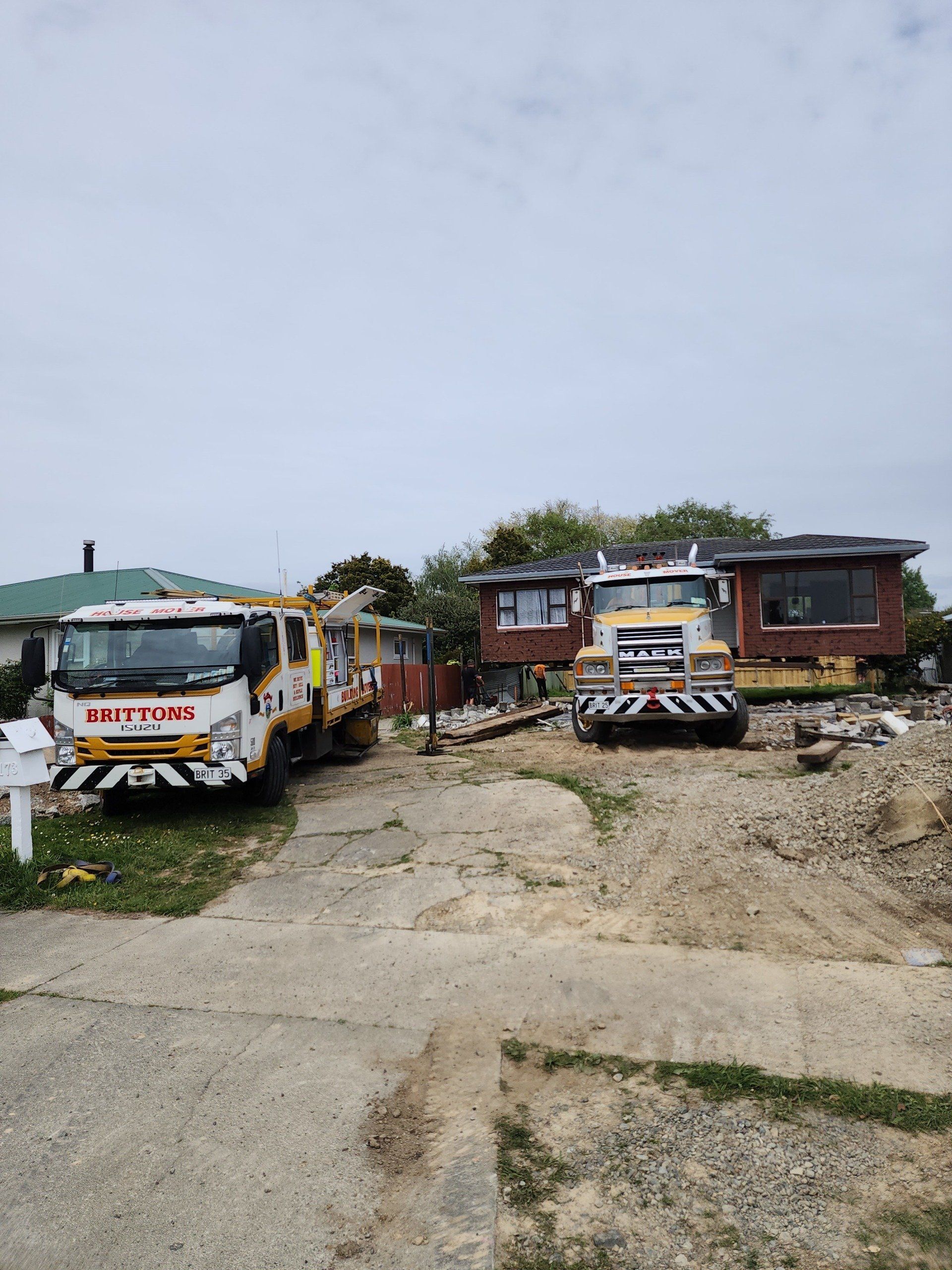 Two yellow utility trucks parked on a cracked driveway beside small houses under a cloudy sky