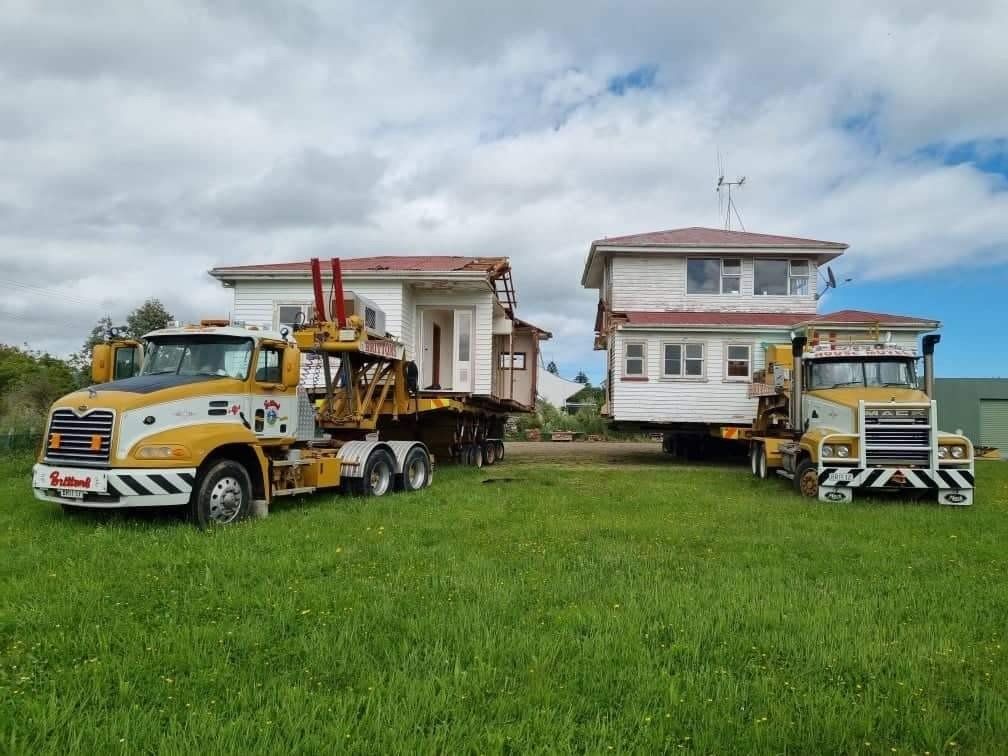 Two yellow tow trucks parked on grass beside a damaged house and a two-story home under cloudy skies