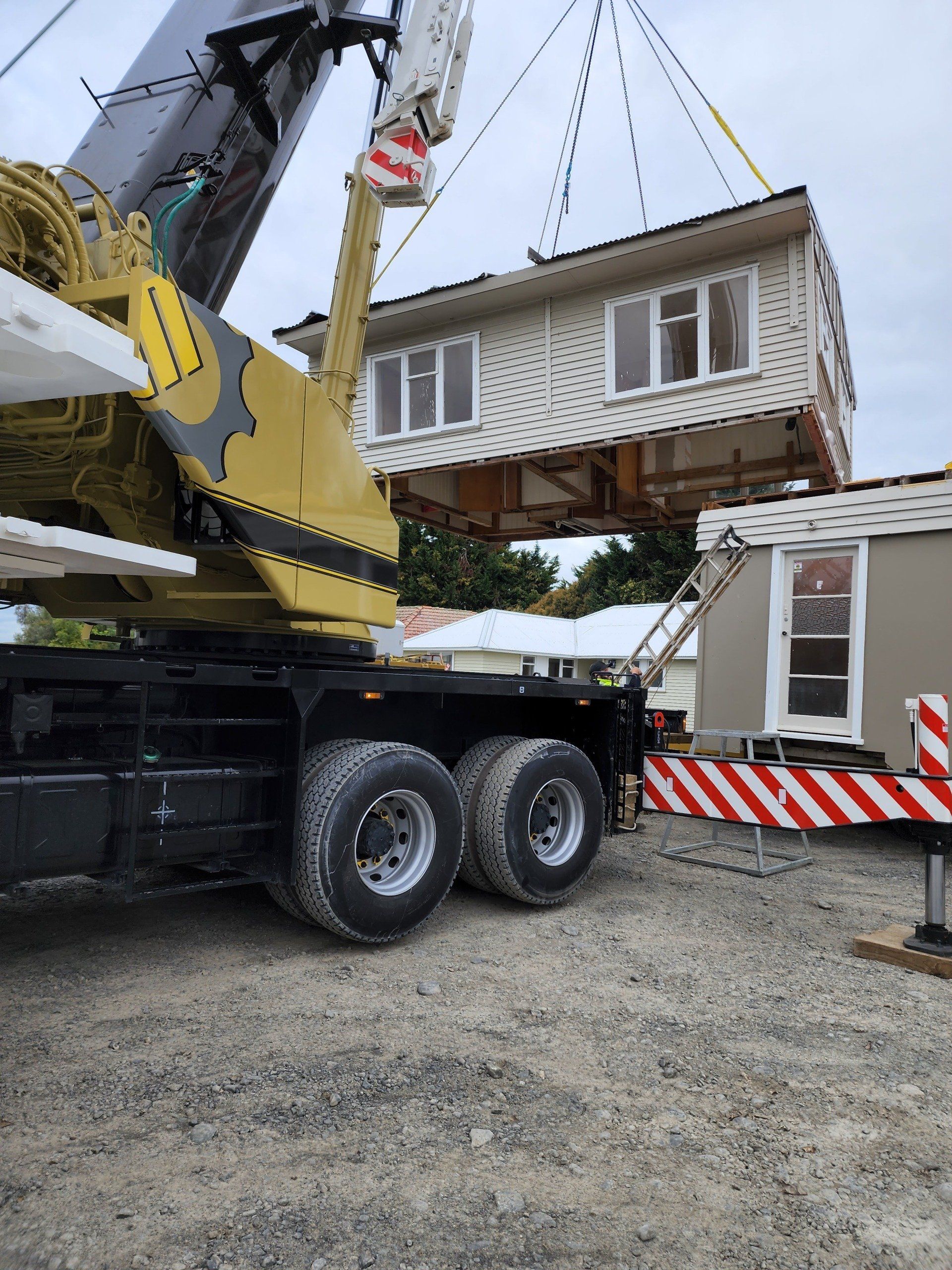 Crane lifting a modular house section beside a red-and-white site trailer on a gravel lot