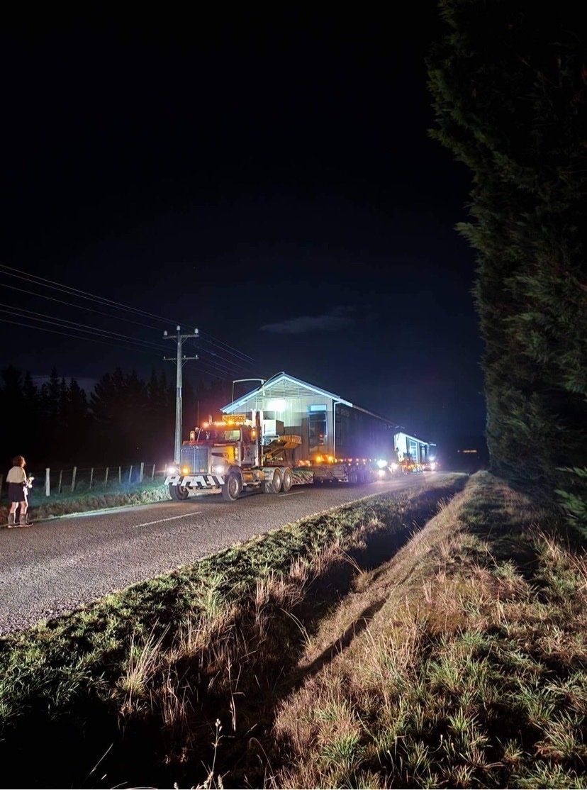 Nighttime road scene with a lit building, vehicles, and people along a rural roadside