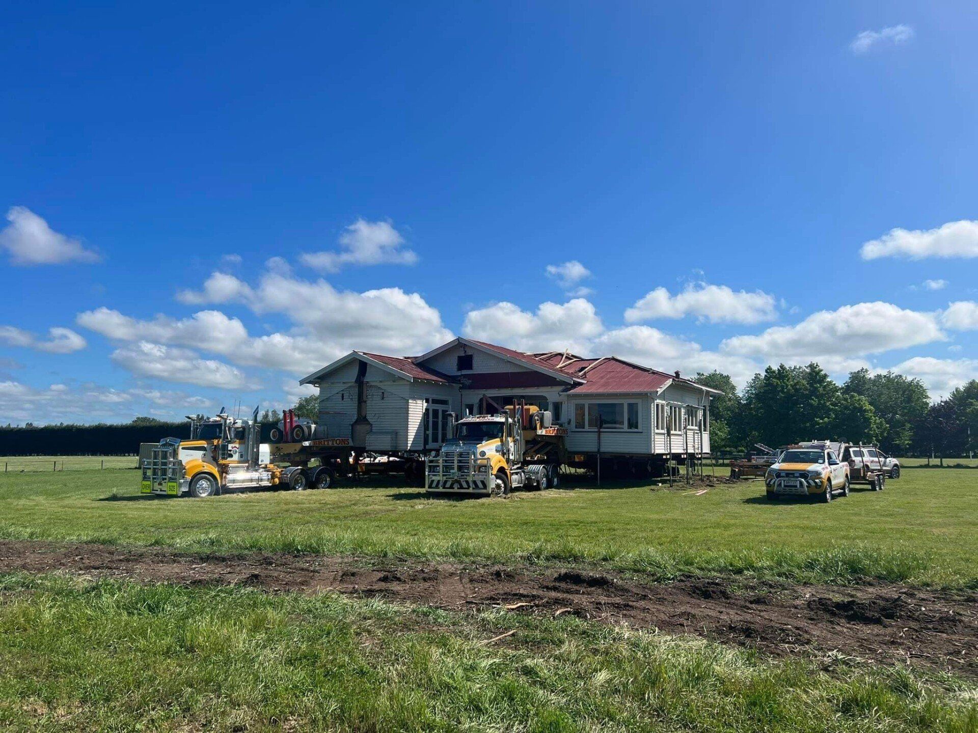 Damaged house being repaired in a grassy field under a blue sky, with construction equipment nearby