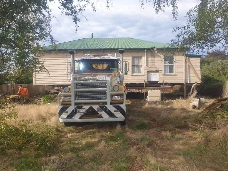 Large truck parked in front of a beige house with a green roof, surrounded by overgrown grass.