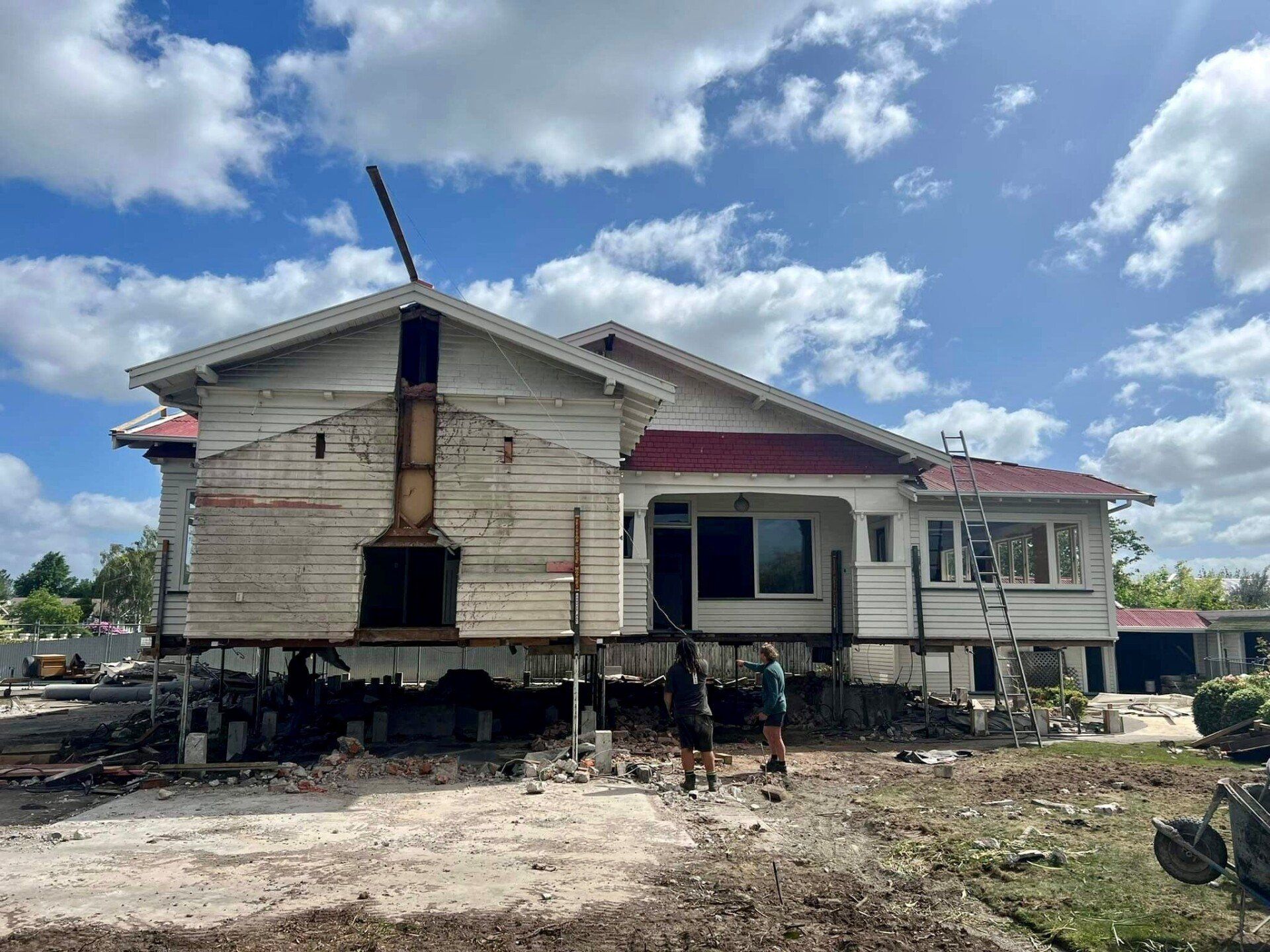 Damaged elevated house with cracked siding and exposed foundation in a muddy yard under a blue sky