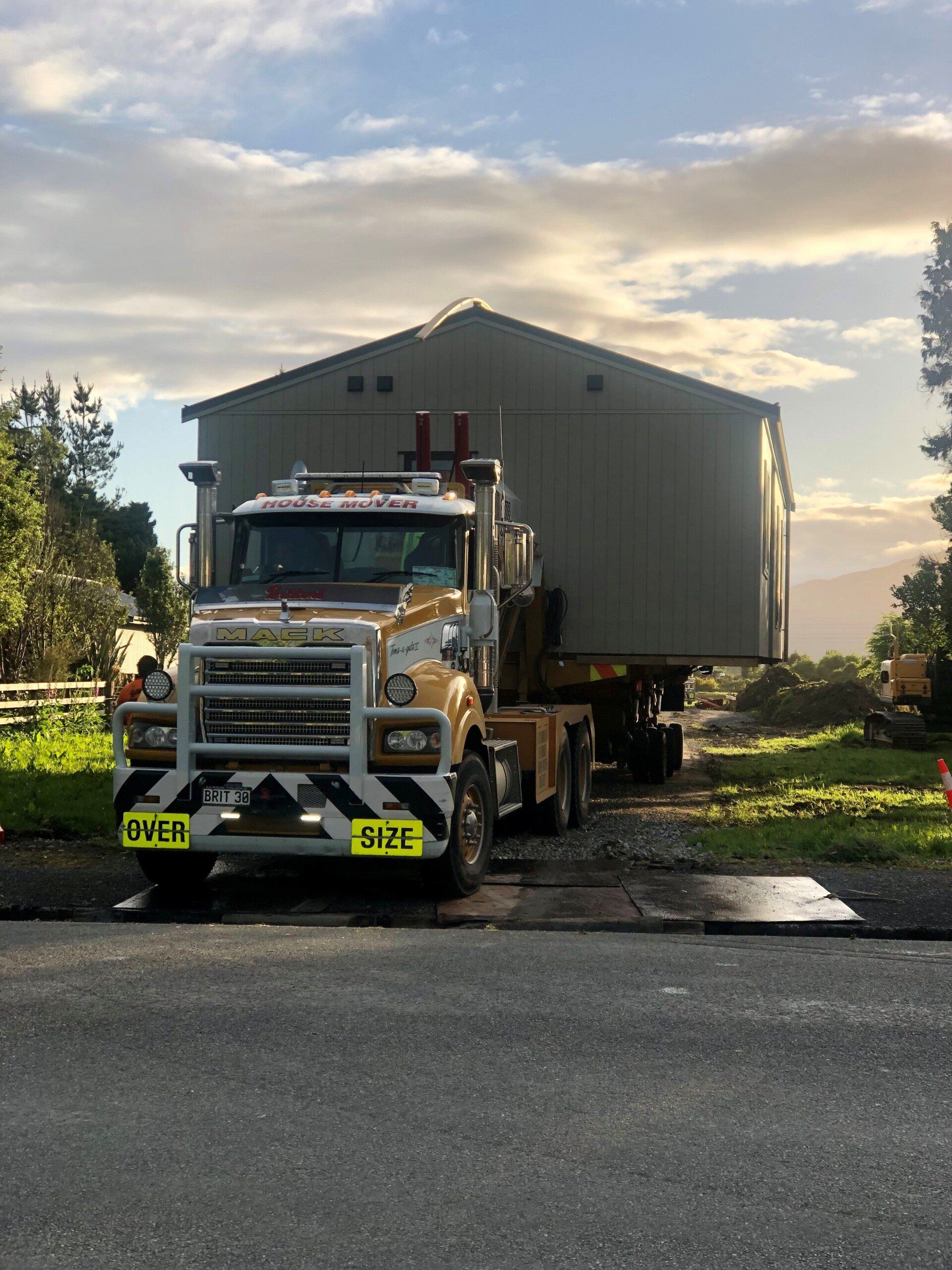 Flatbed truck hauling a gray portable building on a rural road at sunset