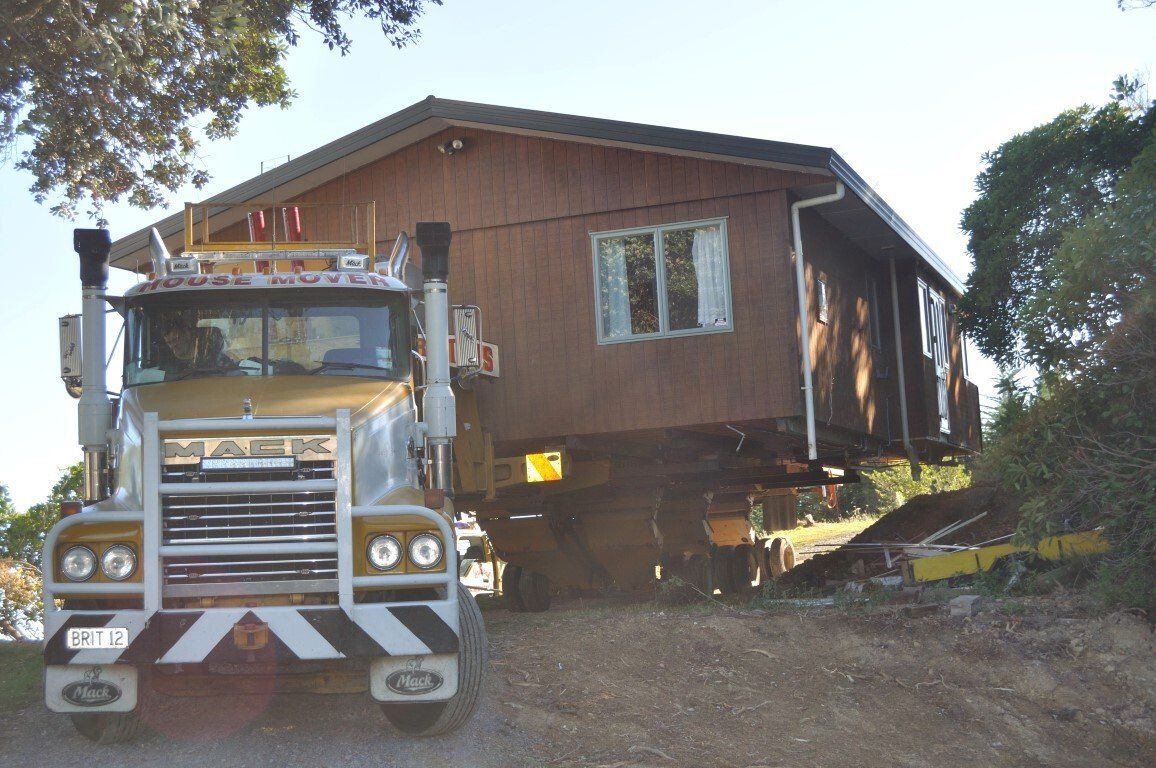 House being lifted by a heavy-duty tow truck on a dirt lot, with trees in the background