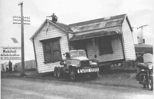 Old wooden house tilted by the roadside with two trucks parked in front and a billboard on the left.