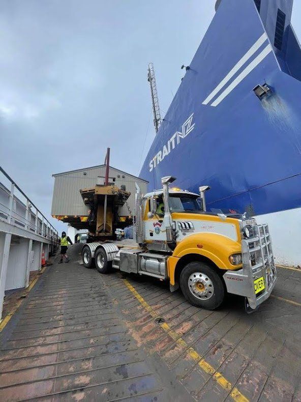 Yellow semi-truck loading cargo at a port beside a large blue ship with a crane