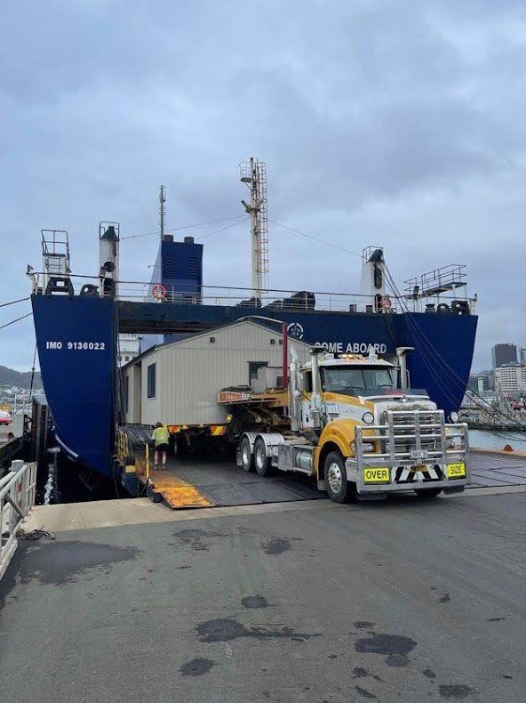 Heavy truck drives onto a blue cargo ship at a dock under a cloudy sky
