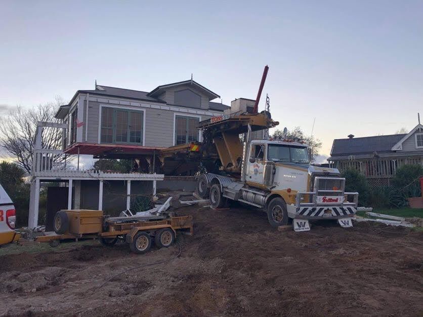 Tow truck lifting a heavy construction vehicle in front of a two-story house, muddy yard at dusk