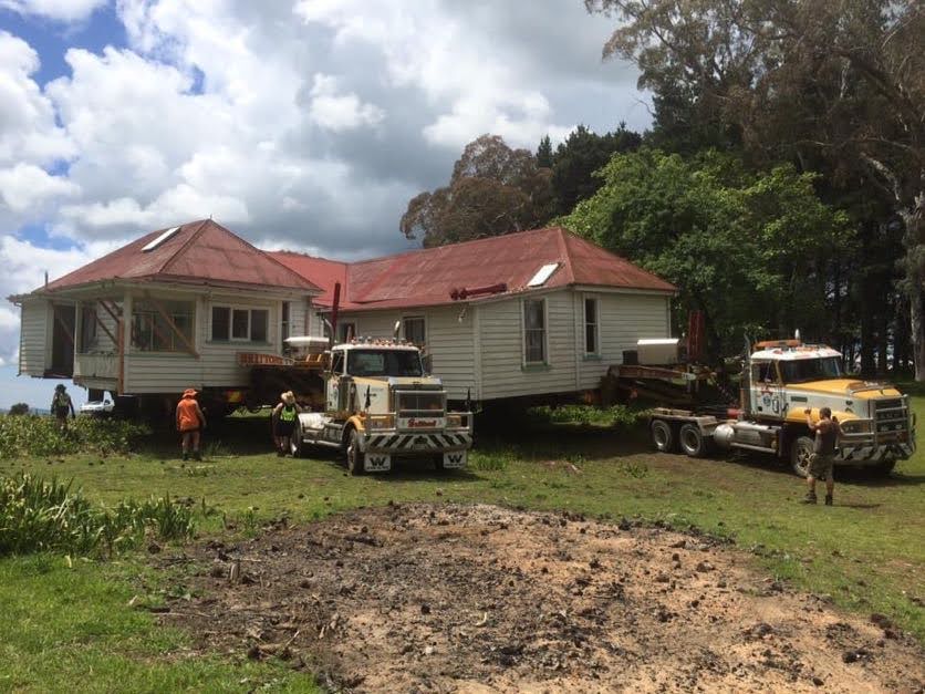 Two trucks and workers move a house in a grassy yard under cloudy skies.