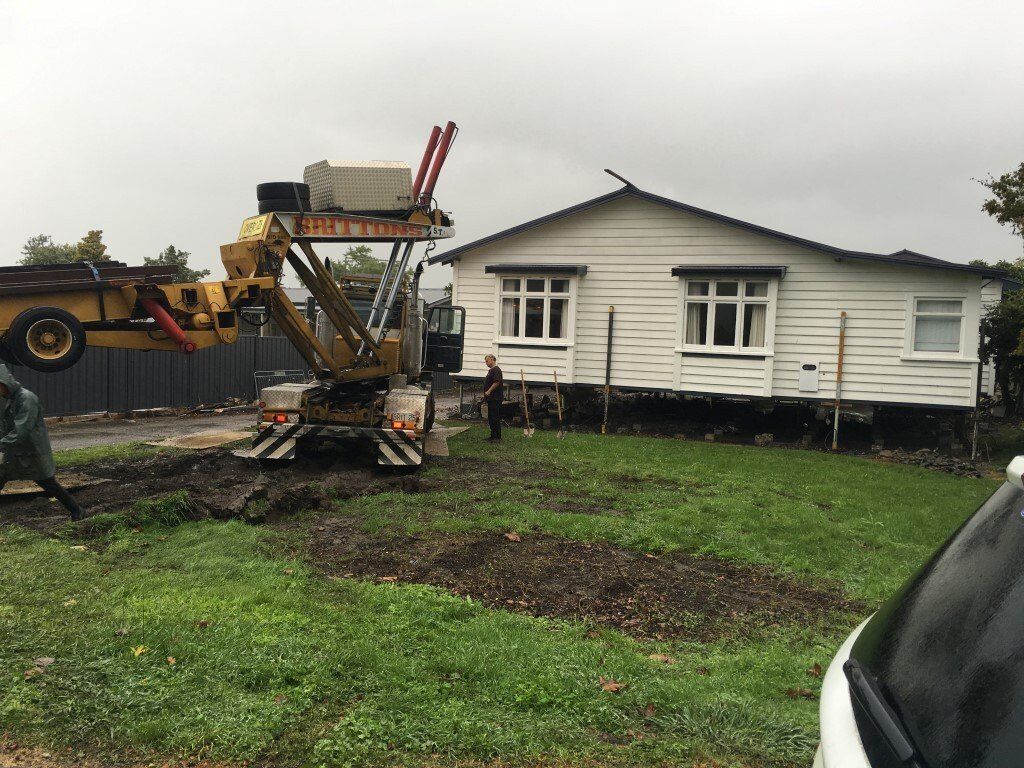 Yellow excavator beside a white house in a muddy yard, lifting debris near the foundation.