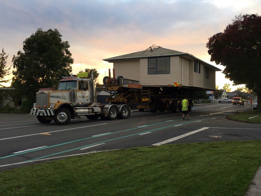 House being transported on a flatbed truck through a street at dusk, with workers nearby