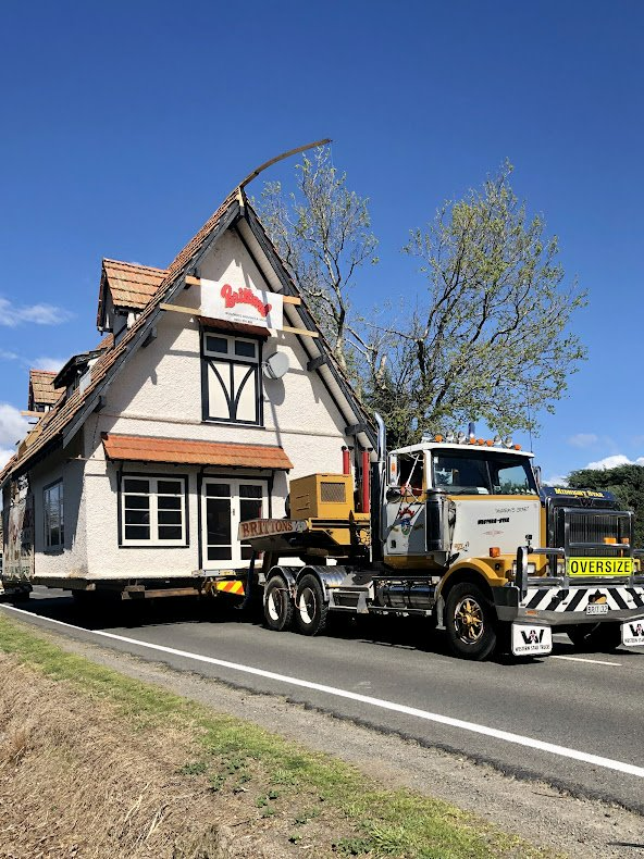 Tow truck lifting a white house-shaped building beside a rural road under a blue sky