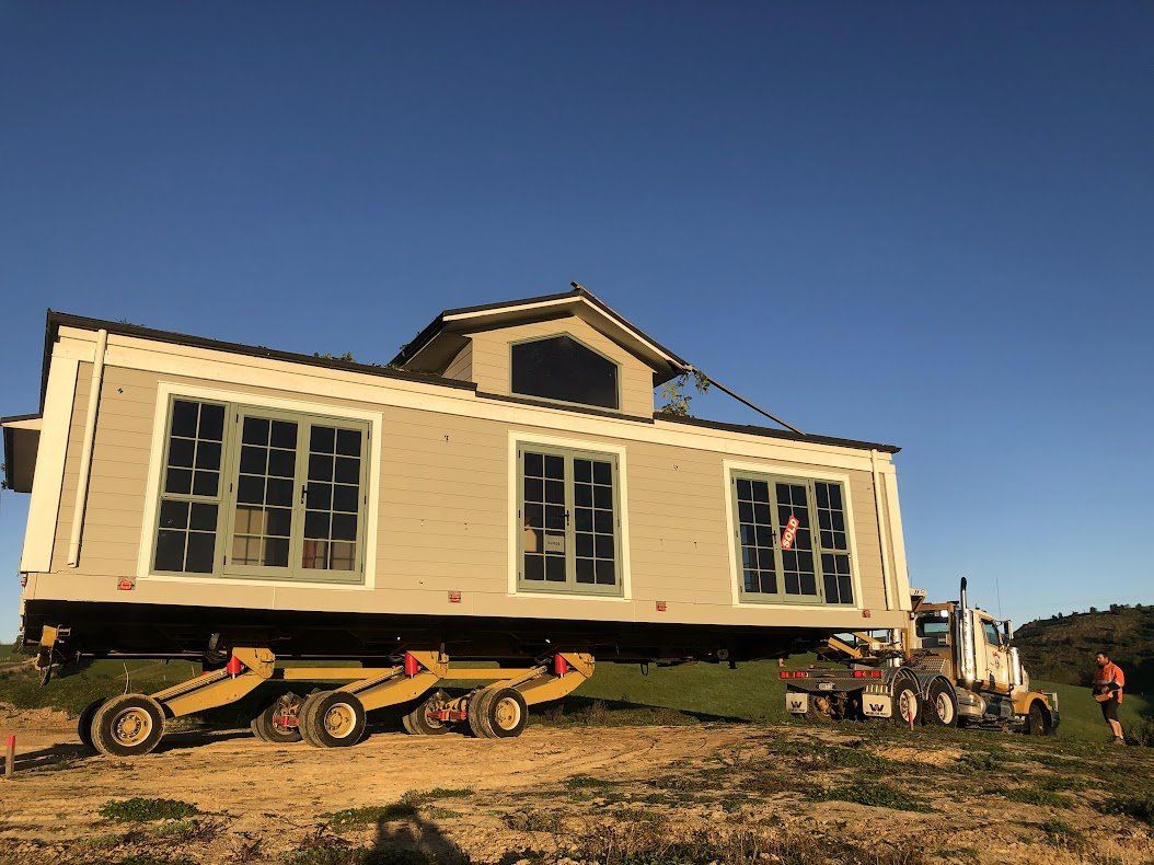 Large cream-colored tiny house being moved on a trailer under a clear blue sky