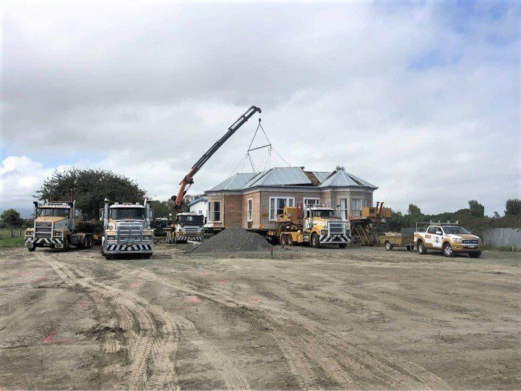 Construction crew lifting roof trusses onto a brick house under construction, with trucks and a crane on a dirt lot.