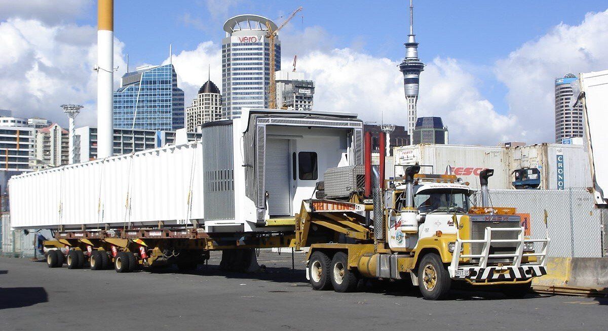 Yellow heavy truck hauling a large white industrial structure in a city harbor, with skyline buildings behind