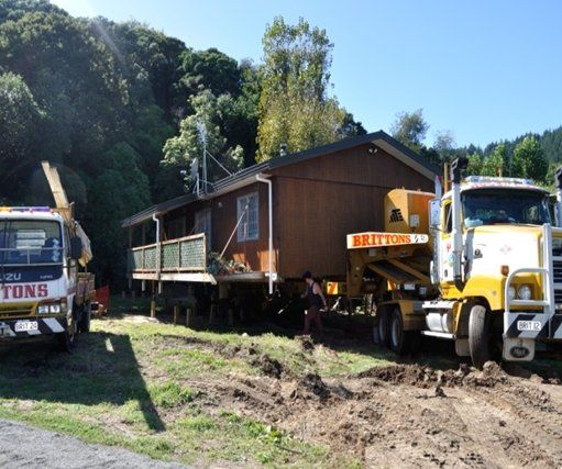 House being transported on a truck, with heavy equipment on a dirt road in a wooded area.