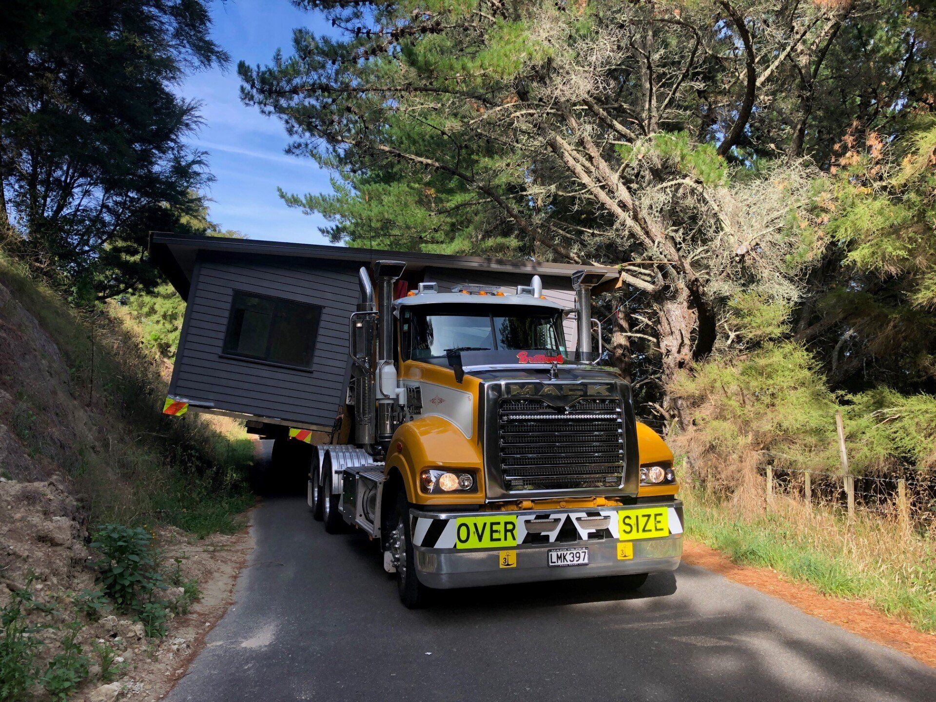Oversize load truck hauling a gray house module on a narrow forest road