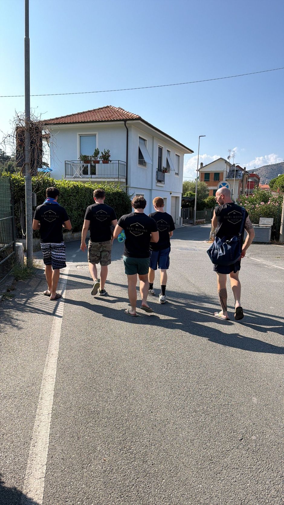 Cinque persone camminano lungo una strada residenziale soleggiata, passando davanti a case bianche